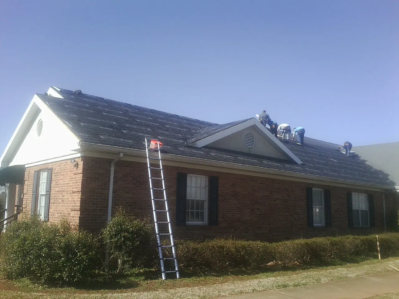 Crew tearing shingles off a brick ranch roof with a ladder set — Emergency Roof Repair work by our crew in Liscomb