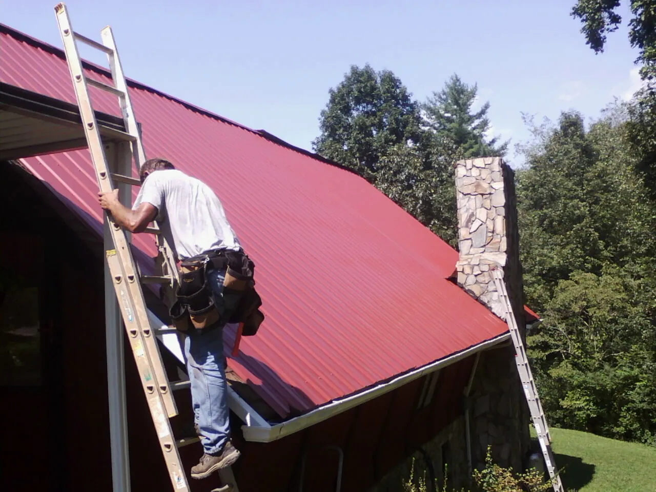 Roofer on a ladder beside a red metal roof and stone chimney — Soffit & Fascia Repair by Roofing Brothers in Liscomb